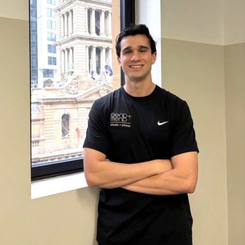 Smiling man in a black athletic shirt with arms crossed, standing by a window in a city office.