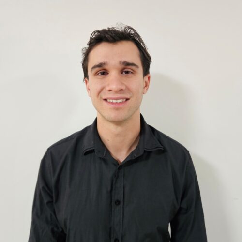 Portrait of a smiling young man with dark hair wearing a black button-down shirt against a light wall.