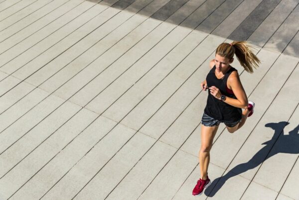 Athlete running on a wide, striped concrete plaza in a black tank top, gray shorts, and red shoes from a top-down view.