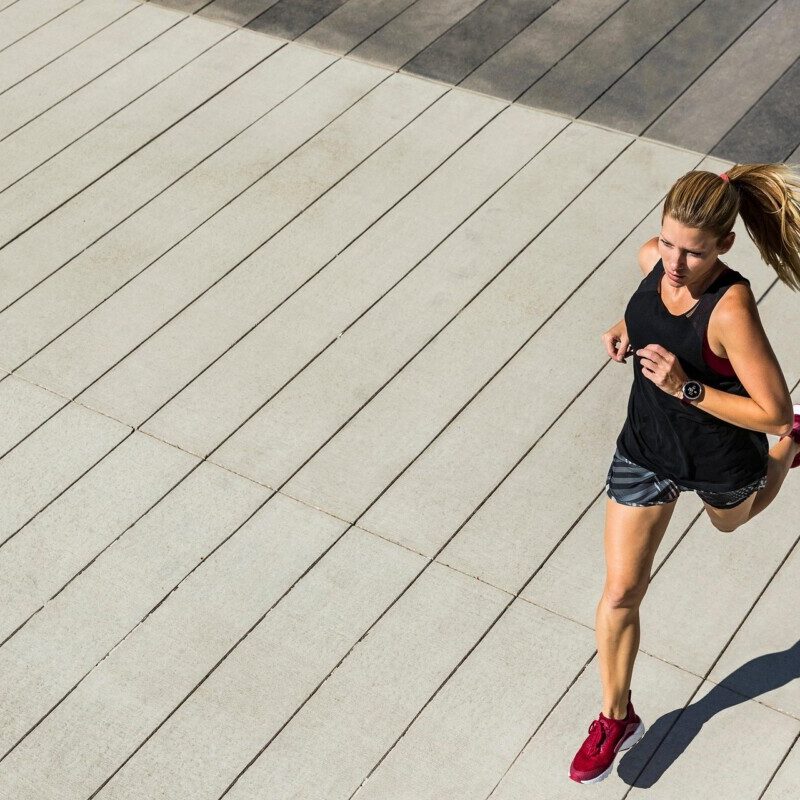 Athlete running on a wide, striped concrete plaza in a black tank top, gray shorts, and red shoes from a top-down view.