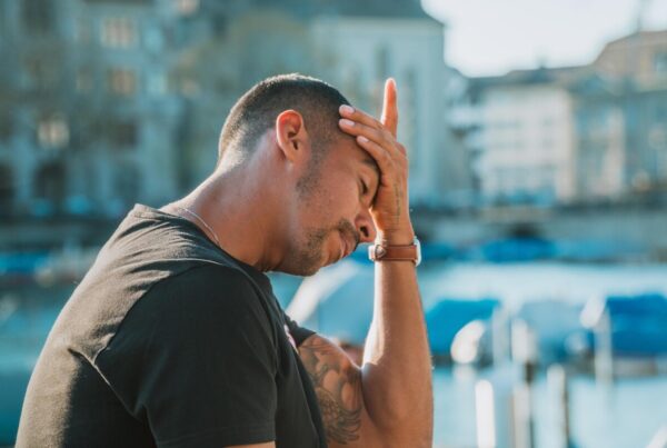 Man in a black t-shirt sits outdoors near a harbor, head in hand, looking stressed or frustrated.