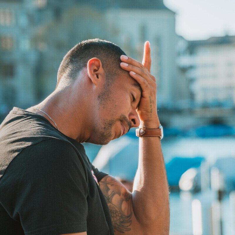 Man in a black t-shirt sits outdoors near a harbor, head in hand, looking stressed or frustrated.