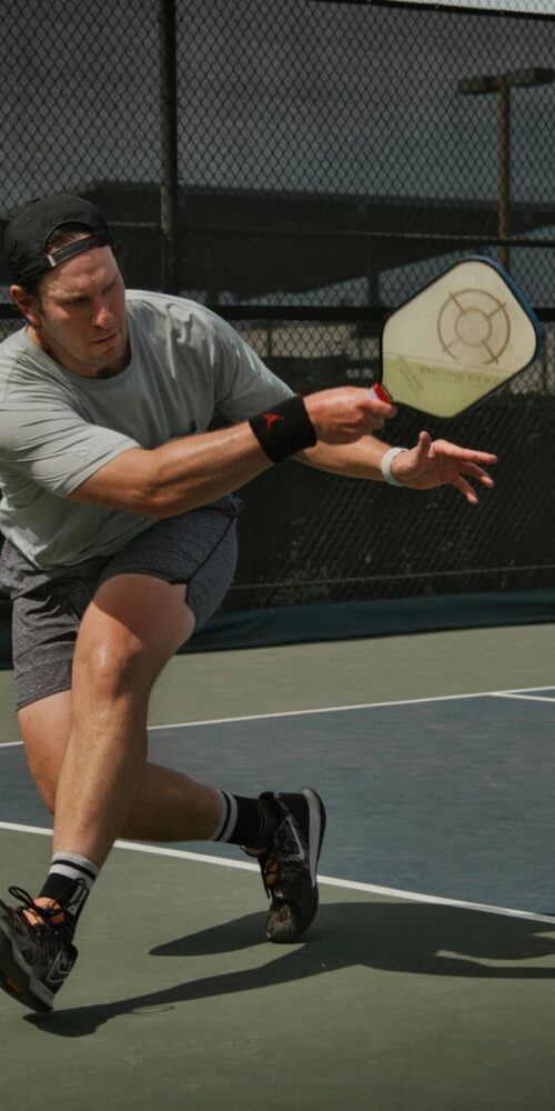 Male athlete in a gray shirt and cap lunges to hit a pickleball with a paddle on an outdoor court fenced backdrop.