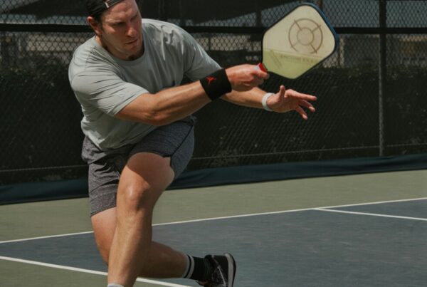 Male athlete in a gray shirt and cap lunges to hit a pickleball with a paddle on an outdoor court fenced backdrop.