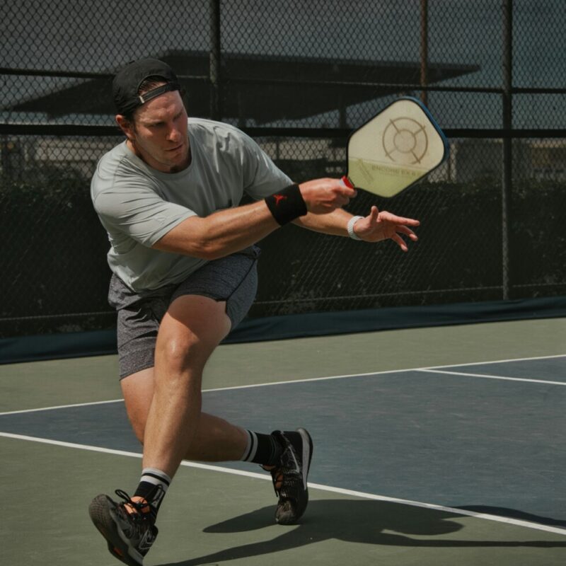 Male athlete in a gray shirt and cap lunges to hit a pickleball with a paddle on an outdoor court fenced backdrop.