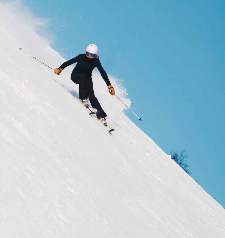 Skier in a black outfit with a white helmet and orange gloves carving down a steep, snowy slope under a clear blue sky, poles in hand.