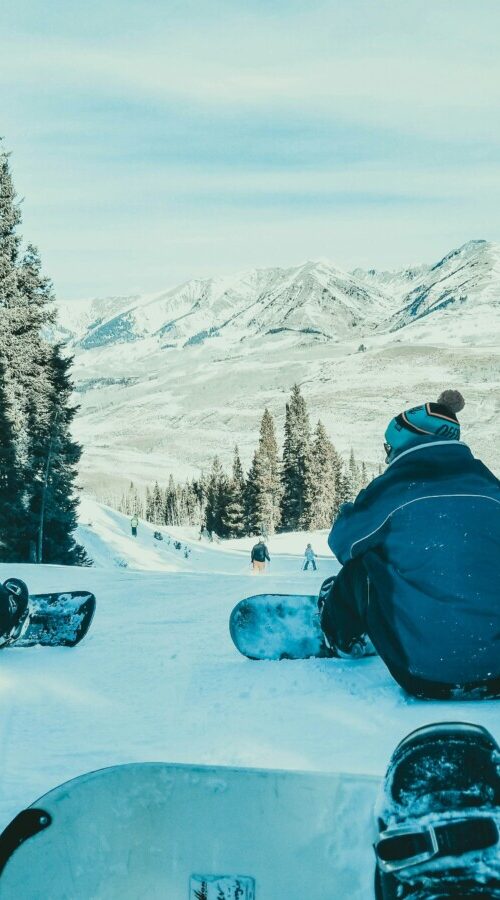 Two snowboarders sit on a snowy slope, facing a forest and distant mountains.