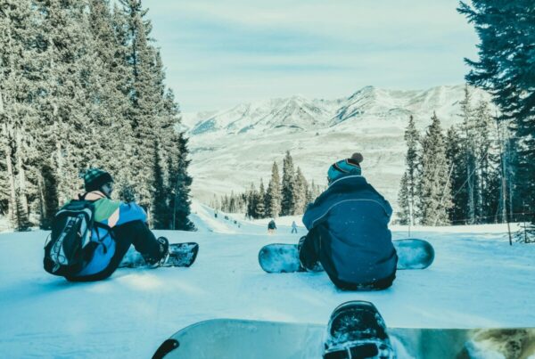 Two snowboarders sit on a snowy slope, facing a forest and distant mountains.