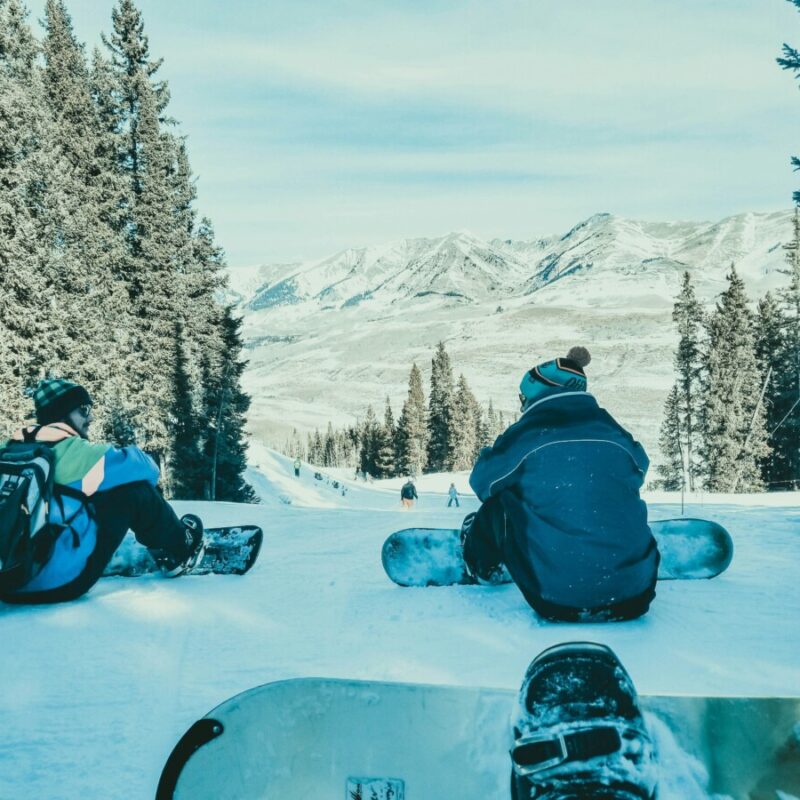 Two snowboarders sit on a snowy slope, facing a forest and distant mountains.
