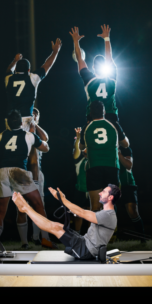 Man doing leg-raise on a Pilates reformer while a rugby team jumps in celebration behind him in a gym. (informative)