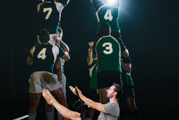 Man doing leg-raise on a Pilates reformer while a rugby team jumps in celebration behind him in a gym. (informative)