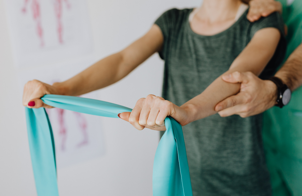 Person holds a turquoise resistance band while a trainer guides their arm during a rehab exercise in a clinical setting.
