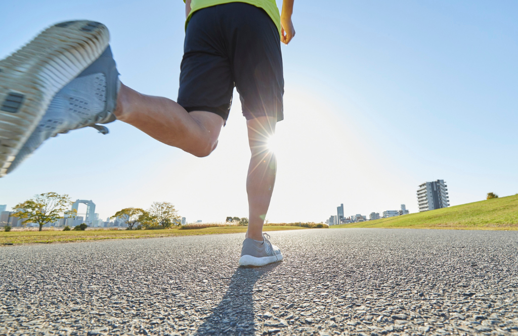 Low-angle shot of a runner's feet in mid-stride on a sunny paved path, city skyline in the distance.