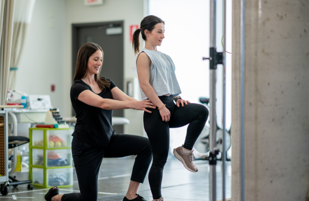 Trainer assists a client as she lifts one leg on a Pilates reformer in a gym setting.