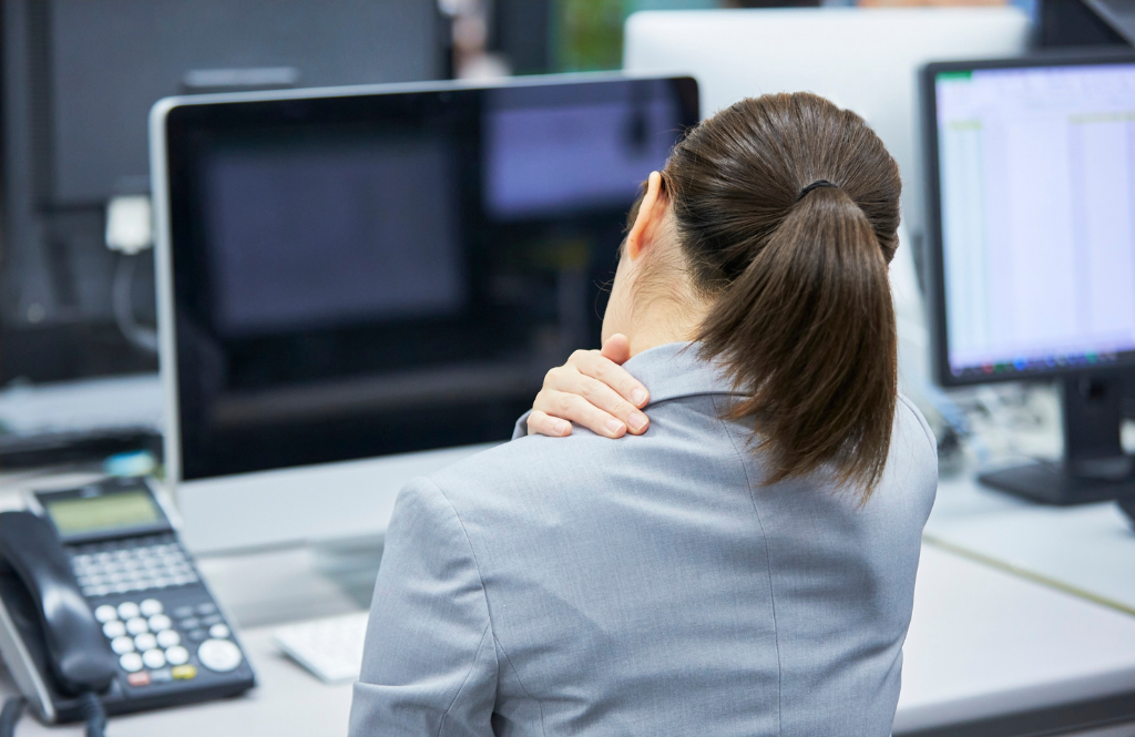 Back view of a person in a gray blazer rubbing their neck at a desk with two computer monitors in front of them.