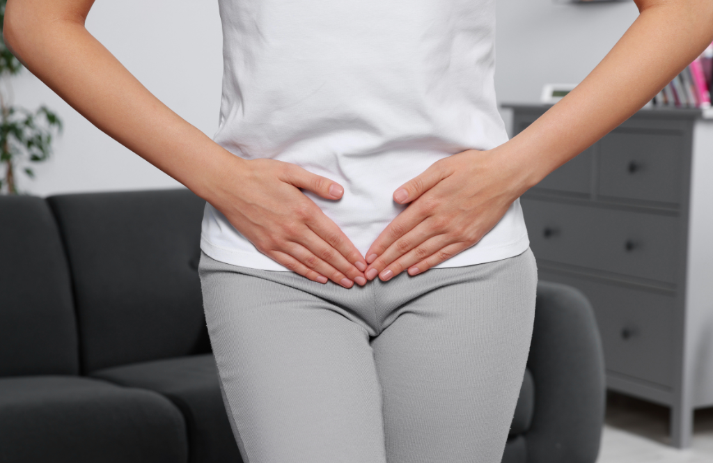 Person standing indoors with hands on the lower abdomen, suggesting stomach discomfort or cramps in a living room setting.